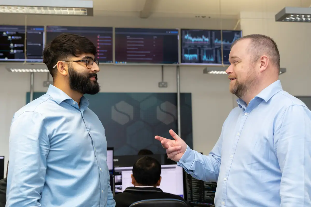 Two IT professionals in a modern office discussing work, with multiple monitors displaying analytics and data in the background