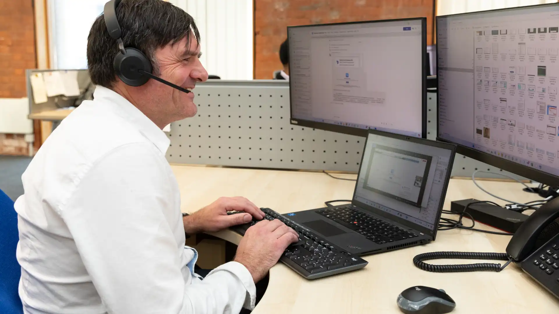 A man wearing a headset, smiling while working on a laptop with multiple monitors displaying IT