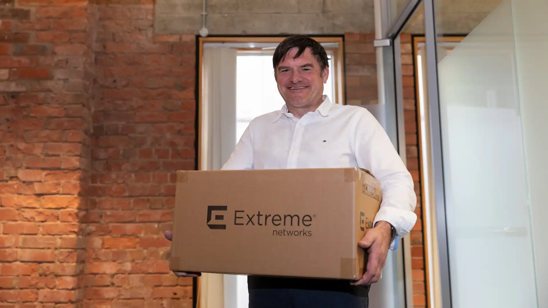 A smiling man in a white shirt holding a cardboard box with the Extreme Networks logo in a modern office