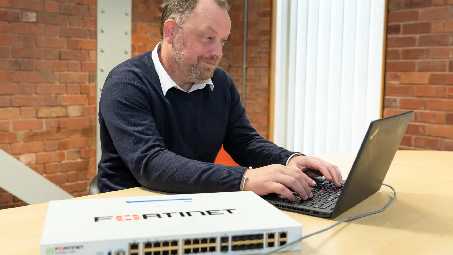 IT professional configuring a network device on a laptop, with a Fortinet switch on the table in a modern office