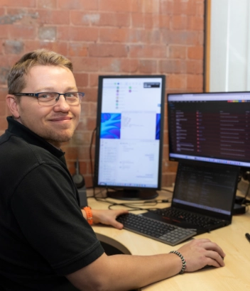 IT professional smiling at his workstation, which features multiple monitors displaying various software applications.