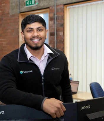 Smiling IT professional wearing a black Syscomm-branded fleece jacket over a white collared shirt