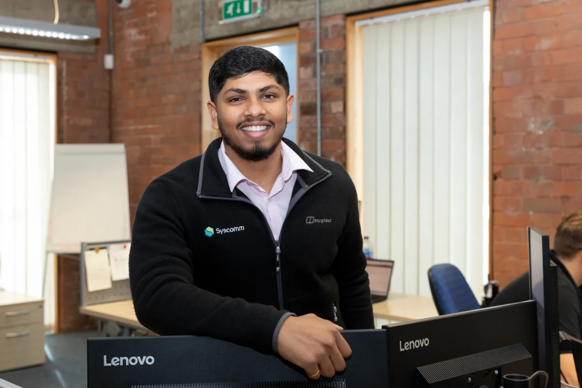 Smiling man standing at a desk in a modern office, wearing a Syscomm-branded jacket landscape