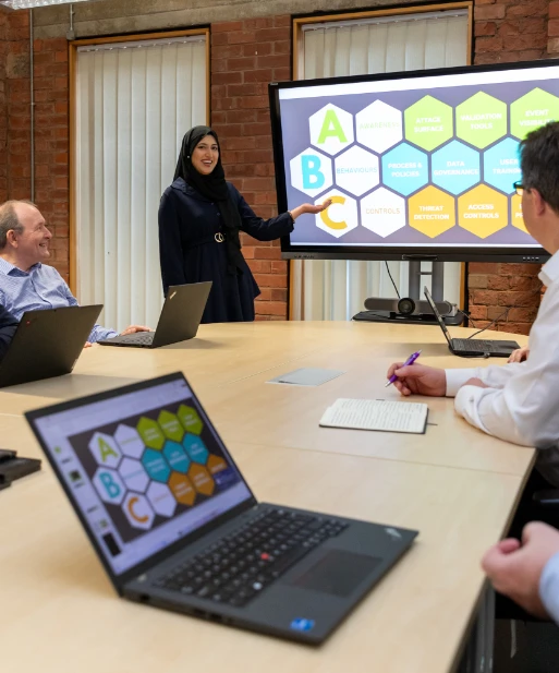 Team members in a meeting room discussing cybersecurity strategies, with a woman presenting a colourful hexagonal diagram on a large screen v2
