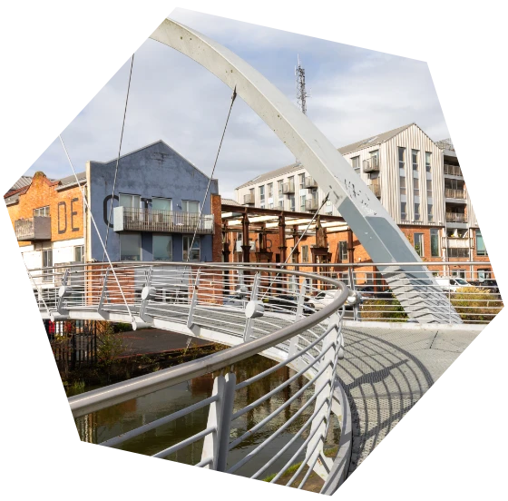A curved pedestrian bridge with metal railings leading towards a modern residential and office area framed in hex