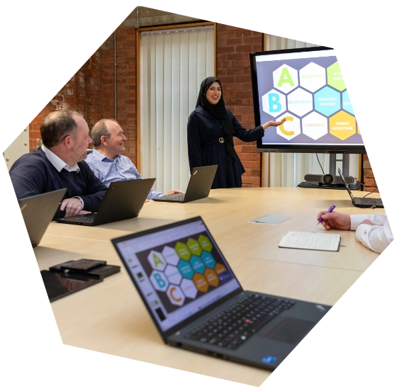 Giving a presentation to a group of colleagues in a modern meeting room, with laptops open and a screen displaying a colourful ABC-themed diagram