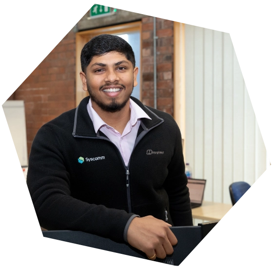 Smiling man in branded Syscomm and DrayTek jacket standing in a modern office environment