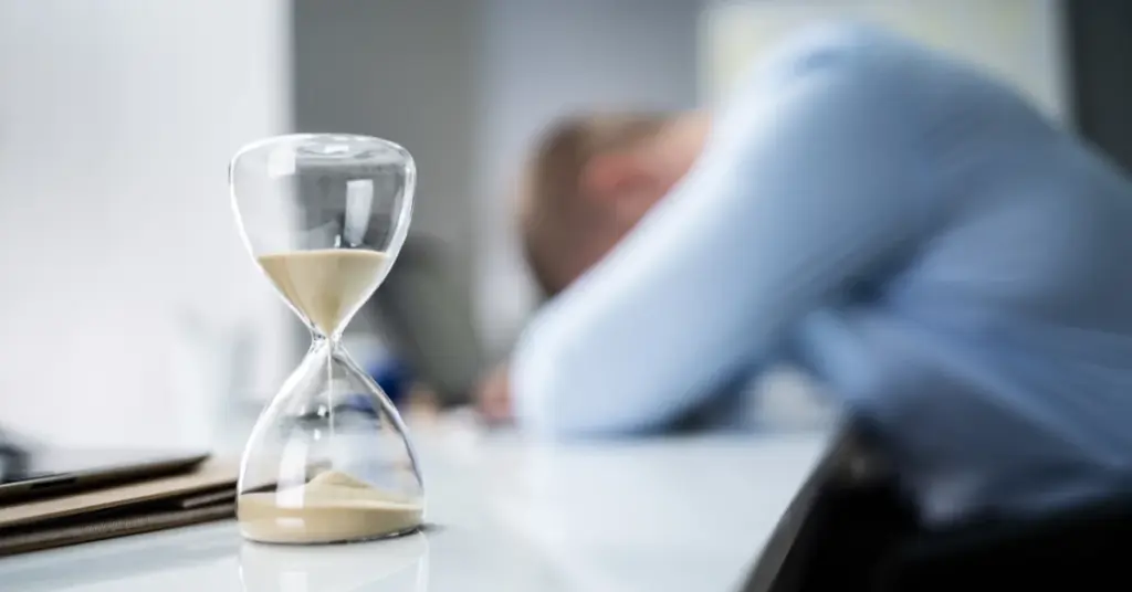 Hourglass running out on a desk beside an exhausted worker resting their head on their arms