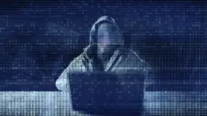 Hooded figure at a laptop with a faint face, overlaid by glitching binary code in a dark blue digital scene.